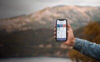 Erfassung von Materialverbrauch mit der Wrocklage App. Hand hält Smartphone mit geöffneter App zur Erfassung von Materialverbrauch, im Hintergrund unscharfe Berglandschaft.