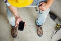 Handwerker auf Baustelle überprüft App auf Smartphone, Bohrmaschine neben seinen Füßen