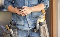 Handwerker in Jeanskleidung tippt auf Smartphone, Werkzeuggürtel mit Reparaturwerkzeugen im Vordergrund