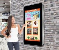 Frau zeigt auf einen an der Wand montierten digitalen Bildschirm mit Werbung für Orange Coffee.