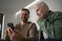 Zwei Männer lächeln und schauen gemeinsam auf ein Smartphone, Symbol für eine App für Pflege- und Sozialdienste zur einfachen Kommunikation und Organisation.