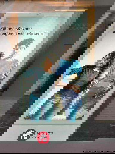 Digitale Werbestele in einer Fußgängerzone zeigt ein Paar mit Umzugskartons und dem Text „Träumen Sie von den eigenen vier Wänden?“. Unten das Logo von Jack Pott Immobilien. Beispiel für digitale Immobilienwerbung im urbanen Raum.