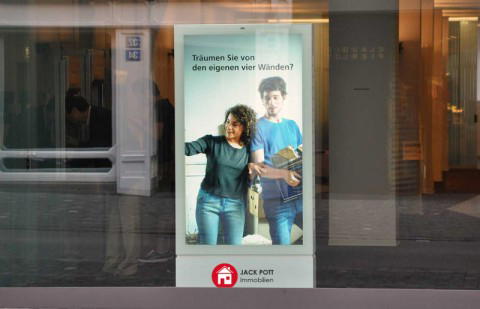 Digitale Außenwerbung für Jack Pott Immobilien. Aufmerksamkeit durch moderne City-Light-Anzeige. Digitale Werbestele in einer Fußgängerzone zeigt ein Paar mit Umzugskartons und dem Text „Träumen Sie von den eigenen vier Wänden?“. Unten das Logo von Jack Pott Immobilien. Beispiel für digitale Immobilienwerbung im urbanen Raum.