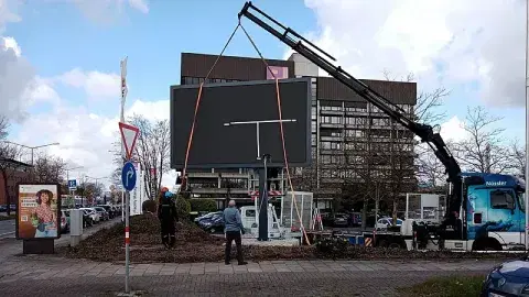 Bauarbeiten an einer digitalen Werbetafel neben einem Parkplatz mit Gehweg im Vordergrund.