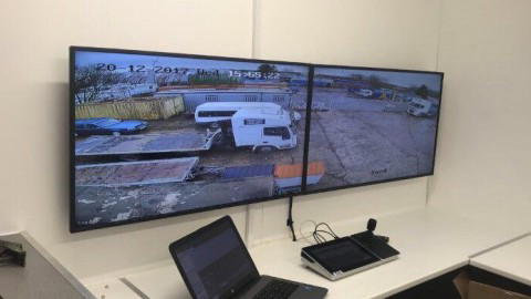 Kontrollraum mit zwei großen Monitoren, die Überwachungsaufnahmen eines Parkplatzes zeigen, Laptop daneben