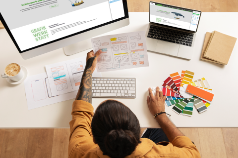 Designer am Schreibtisch mit Farbmustern, Skizzen und Computer beim Erstellen von Webdesign-Layouts